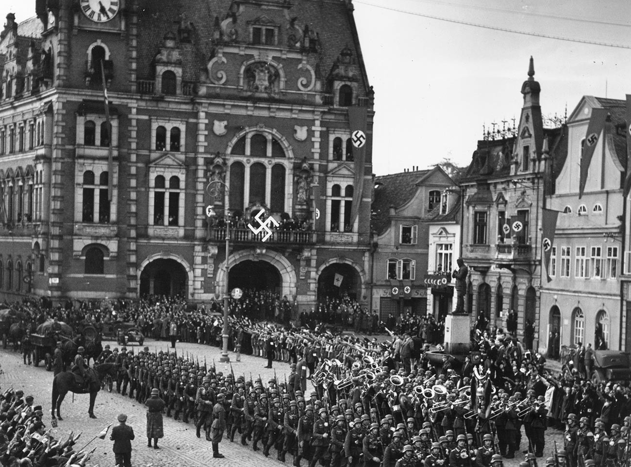 Deutsche Truppen marschieren durch tschechoslowakisches Gebiet auf dem mit Hakenkreuzbannern geschmückten Ramburger Stadtplatz.