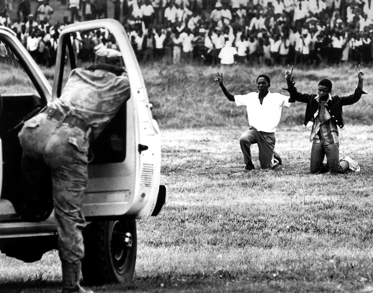 Jovens da cidade de Soweto ajoelhados em frente à polícia com as mãos para o alto mostrando o símbolo da paz, em junho de 1976