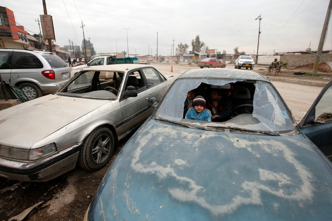 Anak-anak terlihat melalui jendela mobil yang rusak di area industri Mosul, Irak. Sumber: Reuters