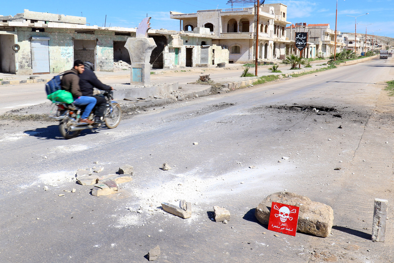 Men ride a motorbike past a hazard sign at a site hit by an airstrike in the town of Khan Sheikhoun in rebel-held Idlib, Syria, April 5, 2017. The hazard sign reads 'Danger, unexploded ammunition.' / Photo: Reuters