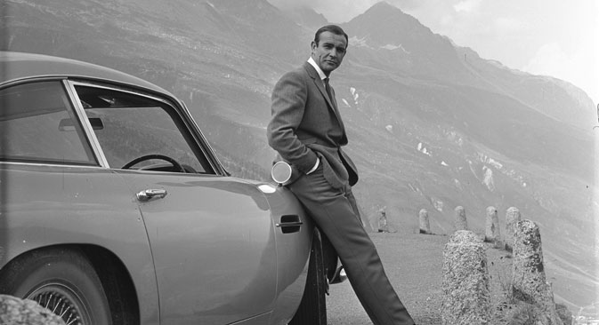 Sean Connery relaxes on the bumper of his Aston Martin DB5 during the filming of location scenes for 'Goldfinger' in the Swiss Alps. Source: Notice - © 1964 Danjaq, LLC and United Artists Corporation.
